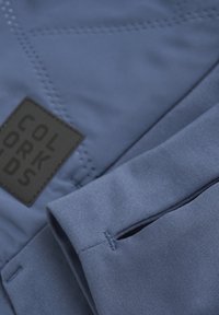 Zbliżenie niebieskiego rękawa z otworem na kciuk obok naszytej czarnej metki marki Colorkids na pikowanej tkaninie.