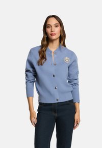 Donna con una cardigan blu chiaro a bottoni con un emblema sul petto e jeans scuri, che sta in piedi contro uno sfondo bianco.