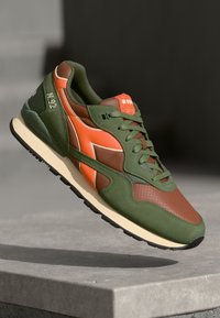 Green and brown sneaker with an orange accent, textured fabric and smooth panels, featuring a cushioned sole and lace-up design.