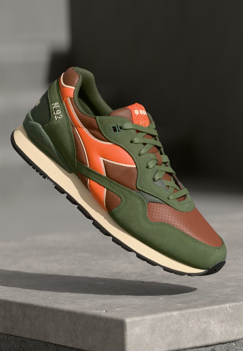 Green and brown sneaker with an orange accent, textured fabric and smooth panels, featuring a cushioned sole and lace-up design.