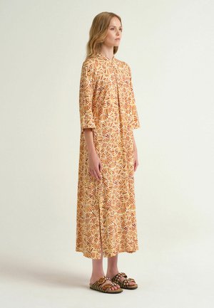 Donna in piedi con un abito midi giallo e arancione a motivo, maniche a tre quarti e sandali con punta aperta su uno sfondo chiaro uniforme.