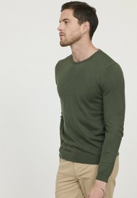 Pull en tricot vert à manches longues, col rond et poignets côtelés. Texture lisse, coupe ajustée, associé à un pantalon beige.