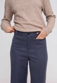 Oseba, oblečena v bež pulover in temno modre hlače s pinstripes visokega pasu, si roke zatakne ali prilagaja pas.