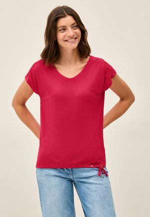 Rotes Kurzarmoberteil mit V-Ausschnitt, aus weichem Stoff. Verfügt über seitliche Versteller mit Bindebändern und kleinen metallischen Akzenten. Getragen mit hellblauen Jeans.