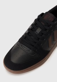 Sorte atletiske sko med en glatt lærtå, semskede detaljer og en teksturert gummisåle. Har svarte lisser og en brun logo-detalj.