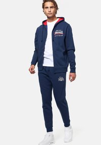 Felpa con cappuccio blu navy con interno rosso, chiusura con zip e logo frontale. Abbinata a pantaloni jogger blu navy con logo e polsini a coste.