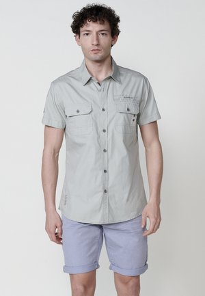 Camisa de botones de manga corta, color gris claro, con dos bolsillos frontales, detalles en negro y un corte relajado. Combinada con pantalones cortos de lavanda arremangados.
