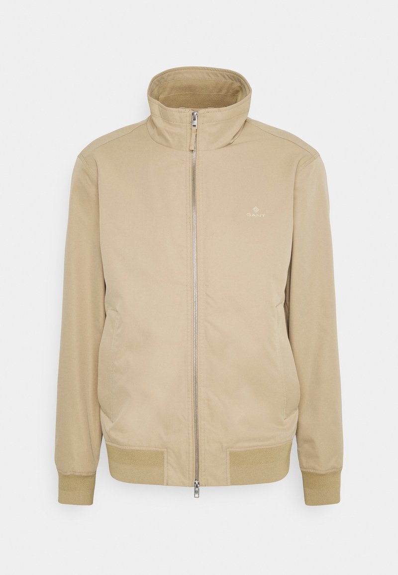 Beige bunda s vysokým límcem, předním zipem a vroubkovanými manžetami. Obsahuje boční kapsy a decentní značkování na hrudi. Hladká textura.