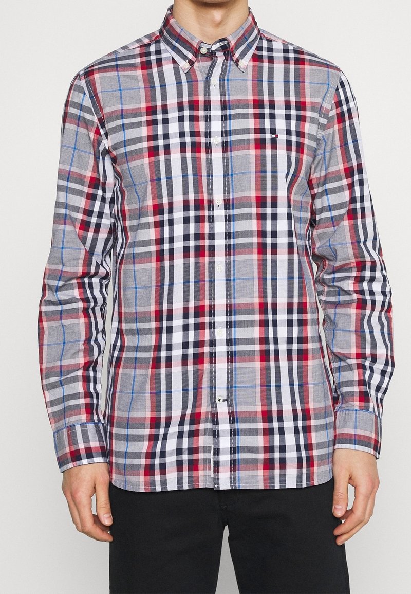 Homme portant une chemise à manches longues boutonnée avec un motif à carreaux rouge, noir, blanc et bleu, associée à un pantalon noir, debout devant un fond uni.