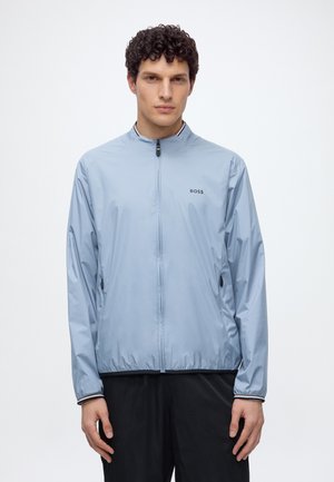 Homme aux cheveux bouclés portant une veste zippée bleu clair avec le logo BOSS et un pantalon noir, debout devant un fond blanc uni.