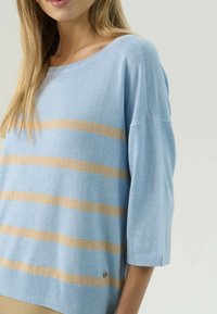 Maglione azzurro chiaro con righe verticali beige, caratterizzato da un ampio scollo, maniche a 3/4 e un piccolo logo in evidenza sull'orlo. Texture morbida.