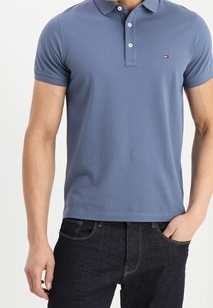 Homme portant un polo bleu ajusté à manches courtes et un jean foncé, une main dans la poche, sur un fond uni.