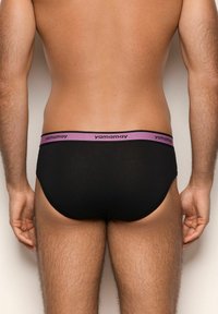 Mann in schwarzen Slips mit einem lila Bund, beschriftet mit "yamamay", steht mit dem Rücken zur Kamera vor einem neutralen Hintergrund.