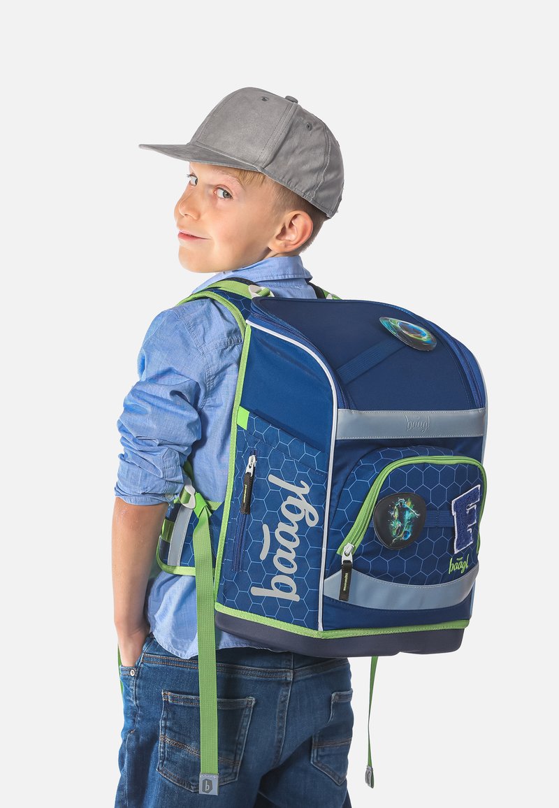 Zaino blu con motivo esagonale, dettagli verdi, cerniere e toppe. Tessuto morbido, forma strutturata. Ragazzo che indossa una maglietta blu e un cappellino.
