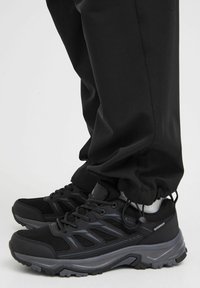 Scarpe sportive nere impermeabili con dettagli in rete, suole in gomma, accenti testurizzati e riflessi grigi, abbinate a pantaloni neri e calze bianche.