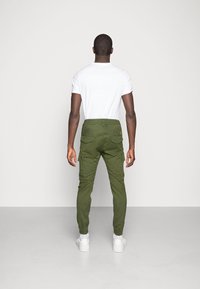 Olivgrüne Cargo-Hosen mit elastischen Bündchen und seitlichen Taschen, kombiniert mit einem weißen T-Shirt und weißen Turnschuhen, von hinten betrachtet.