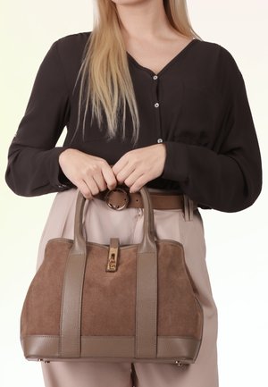 Vrouw in zwarte blouse en beige broek houdt voor zich een bruine suède- en lederen handtas met gouden sluiting.