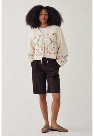 Mujer con cabello rizado y voluminoso lleva un cárdigan crema bordado con flores, top blanco, pantalones cortos oscuros hasta la rodilla y mocasines sin cordones color caoba.