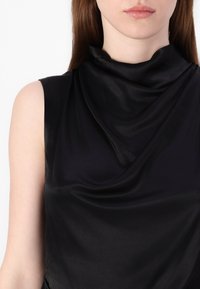 Svarte utenermede topp med en cowl-hals, laget av glatt, glanset stoff. Draperingen skaper myke folder som fremhever teksturen og glansen.