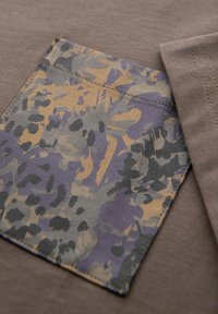 Hnědá látková halenka s prošitým kapsou, která má kamuflážní vzor v odstínech fialové, modré a béžové. Okraje kapsy jsou pečlivě zpracovány.
