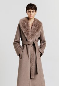 Cappotto marrone con colletto in pelliccia morbida, dettaglio della cintura, maniche lunghe e tasche laterali; tessuto testurizzato con una finitura liscia.