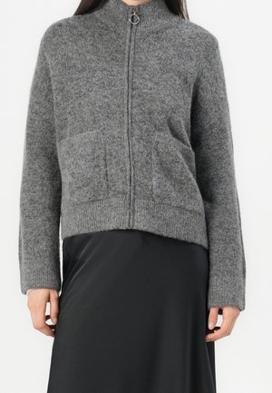 Pull à zip gris avec col montant, poignets côtelés, et deux poches avant. Tissu doux et texturé avec une coupe légèrement raccourcie.