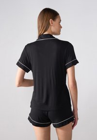 Schwarzes kurzärmliges Top mit Kragen und weißem Piping, kombiniert mit schwarzen Shorts. Der Stoff wirkt weich mit einer glatten Textur.