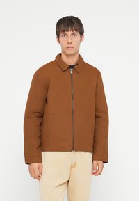 Brown padded jacket with a collared neckline, front zipper, and side pockets, worn over light-colored jeans. Smooth texture, casual design.