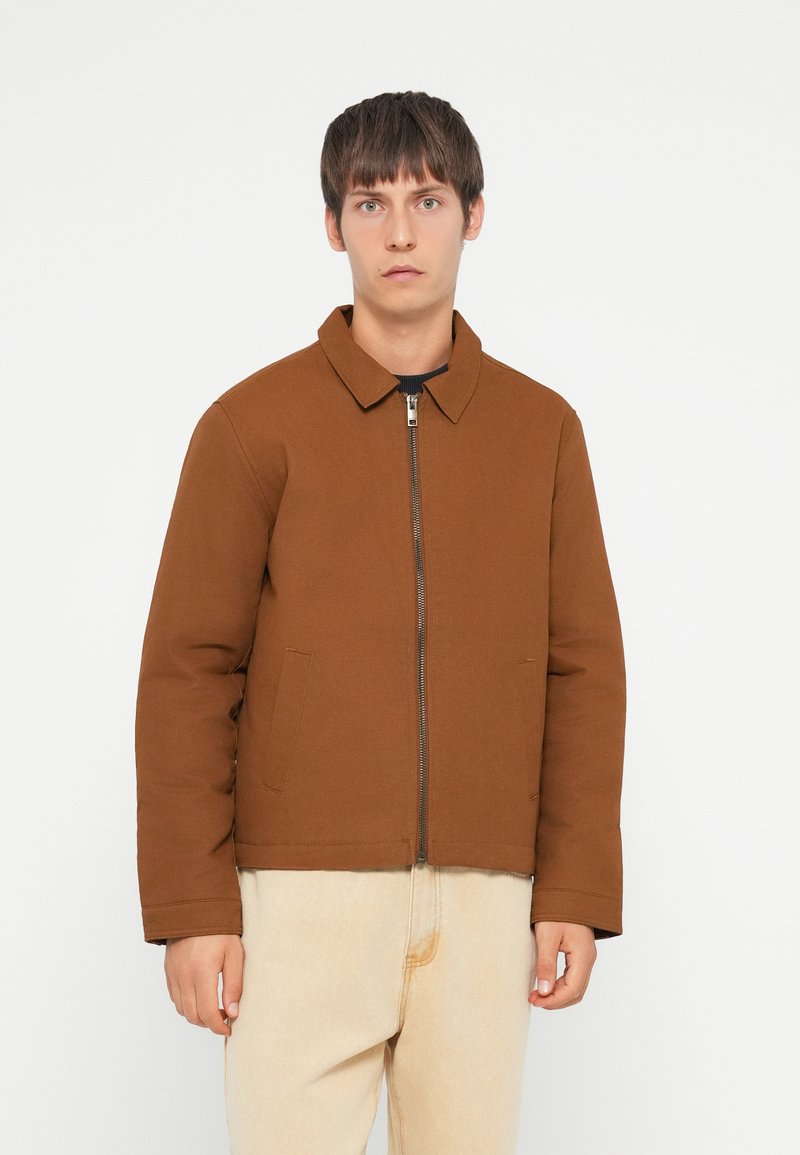 Brown padded jacket with a collared neckline, front zipper, and side pockets, worn over light-colored jeans. Smooth texture, casual design.