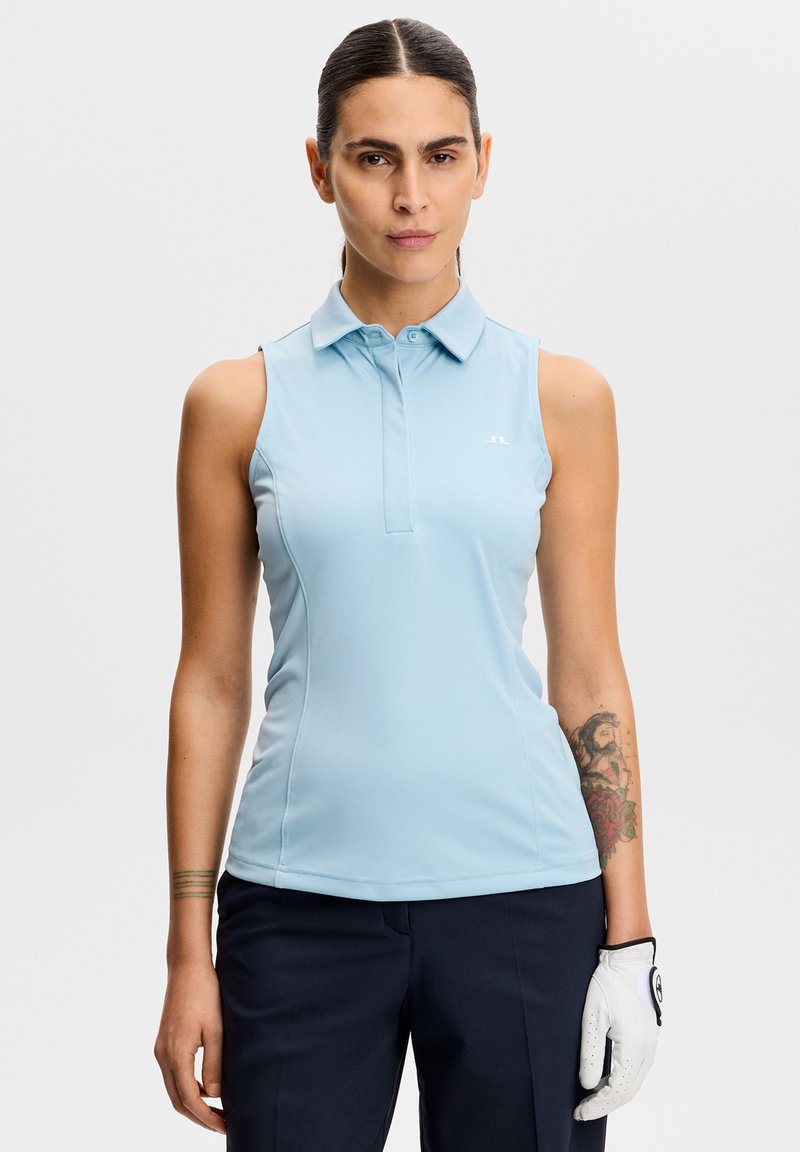 Femme portant un polo sans manches bleu clair et un pantalon bleu marine, avec un tatouage sur le bras gauche et un gant de golf blanc à la main droite.
