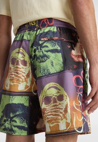 Vícebarevné plavky mají koláž grafických tváří v živé zelené, oranžové a fialové barvě. Elastický pás obsahuje zadní kapsu.