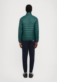 Teal pufferjakke med quiltet design og ståkrage, kombinert med marineblå joggebukser. Modellen står med ryggen til kameraet, og viser detaljer på jakken.