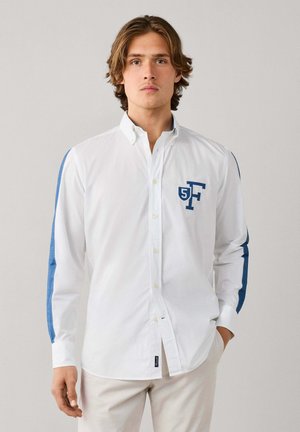 Homme aux cheveux bruns mi-longs porte une chemise blanche à boutons avec des rayures bleues sur les manches et un logo bleu "5F" sur la poitrine, debout immobile.