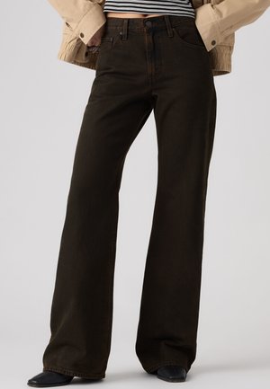 Personne portant un jean taille haute, jambes larges, brun foncé, des chaussures noires, une veste beige et un top court rayé noir et blanc.