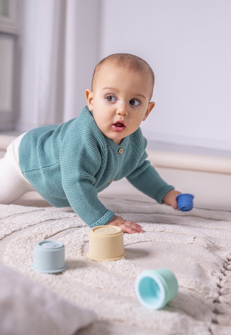 Pull en tricot teal clair avec des boutons, bébé en train de ramper sur un tapis texturé blanc. Trois gobelets empilables doux et colorés disposés à proximité.