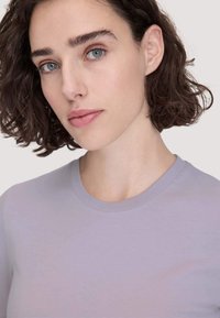 Vrouw met kort, krullend donker haar en blauwe ogen, die een lichtpaarse crew-neck shirt draagt tegen een effen achtergrond.