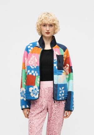 Femme aux cheveux blonds bouclés portant une veste polaire à motifs floraux multicolores sur un haut noir et un pantalon rose à imprimé animal.