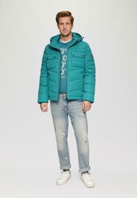 Teal gewatteerde jas met een capuchon en voorzakken, gecombineerd met een blauw graphic t-shirt en vervaagde distressed jeans. Witte sneakers maken de look af.