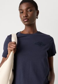 Tricou bleu marin cu mânecă scurtă, cu detalii brodate pe piept. Modelul poartă o geantă bej de umăr, având o textură netedă.