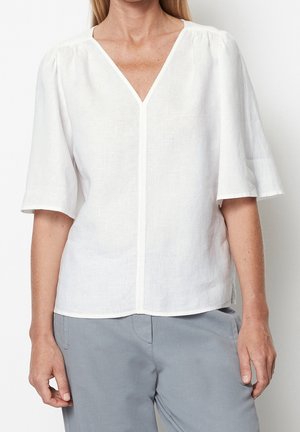 Kvinde iført en hvid V-hals bluse med korte, løstsiddende ærmer og lysegrå bukser, stående foran en ensfarvet baggrund.