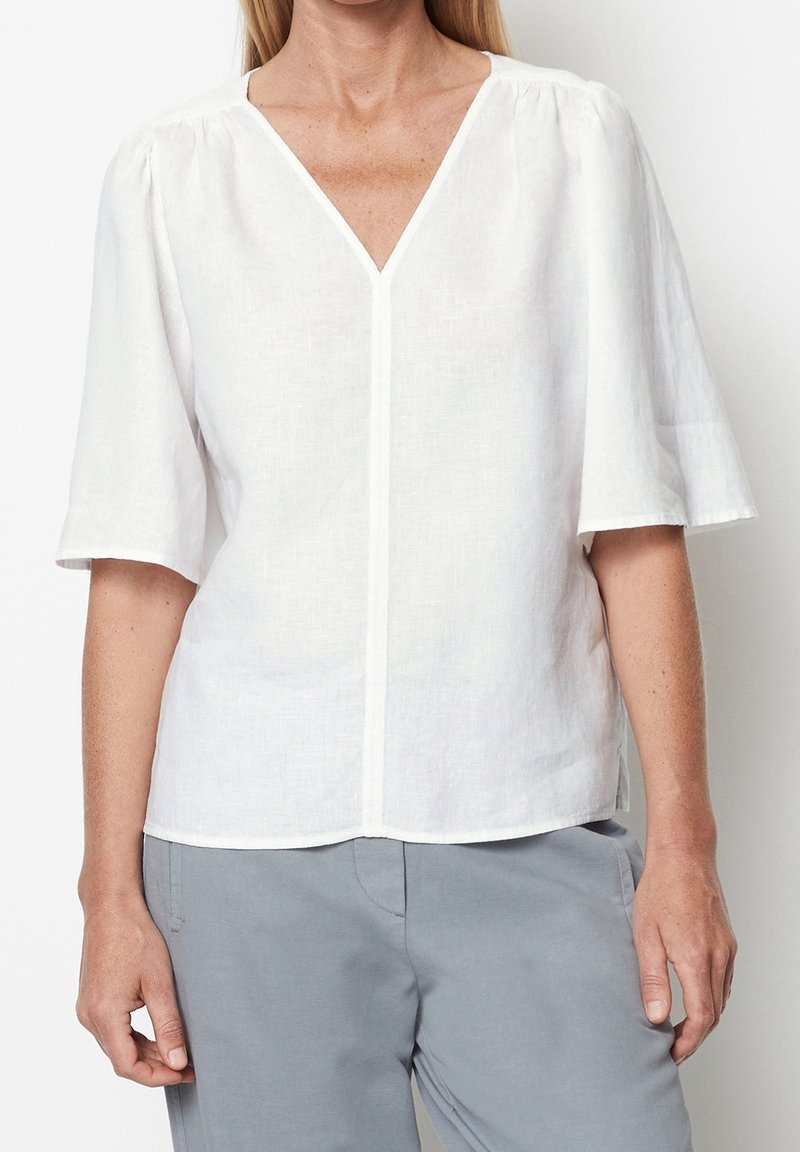 Femme portant un chemisier blanc à col en V avec des manches courtes et amples et un pantalon gris clair, debout devant un fond uni.