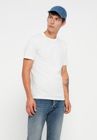 Ung mann iført hvit T-skjorte, blå jeans og blå caps, som ser til siden med en tatovering på venstre underarm.