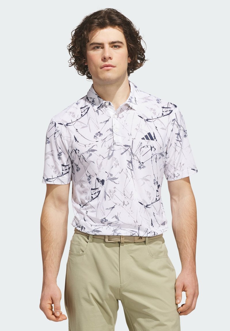Jeune homme aux cheveux bouclés portant un polo blanc à manches courtes avec imprimé floral noir et pantalon beige sur un fond uni.