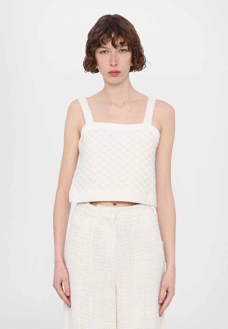 White, textured cropped top with thick, square straps. Notable diamond pattern and straight hem, paired with light, loose-fitting trousers.