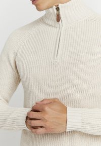 Beige strikkegenser med høy krage og halv glidelås, med teksturert stoff og ribbestrikkede mansjetter. Glidelåsen sikres med en lærpulltab.