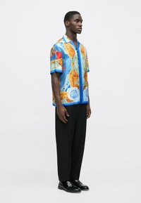 Short-sleeve shirt in blue with multi-colored patterns, paired with black trousers and black shoes. Features a relaxed fit and button closure.