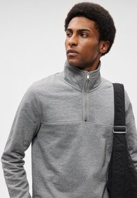 Γκρίζο φλις μισό-λίγο πουλόβερ με ψηλό γιακά, αντίθετη ραφή και λεία υφή. Μια μαύρη τσάντα με λουρί είναι κρεμασμένη στον ώμο.