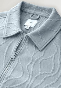 Pull en maille bleu clair à fermeture éclair avec des motifs géométriques texturés, un col, et une étiquette blanche visible indiquant la taille et la marque "NEXT" à l'intérieur.
