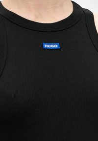 Detail čierneho rebrovaného tielka s malou modrou obdĺžnikovou nálepkou označenou „HUGO“ v strede pri výstrihu.