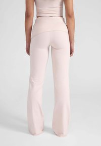 Vrouw in hoge taille, lichtroze flared broeken en een bijpassende korte top, staande met ontspannen handen langs haar zij.